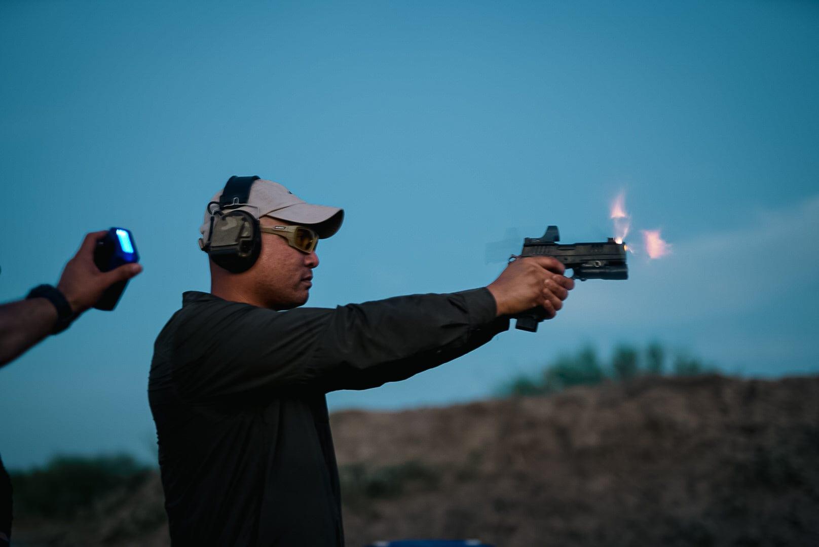 A shooter firing a red-dot pistol at dusk, muzzle flash lit against a blue sky, an instructor timing the string beside him