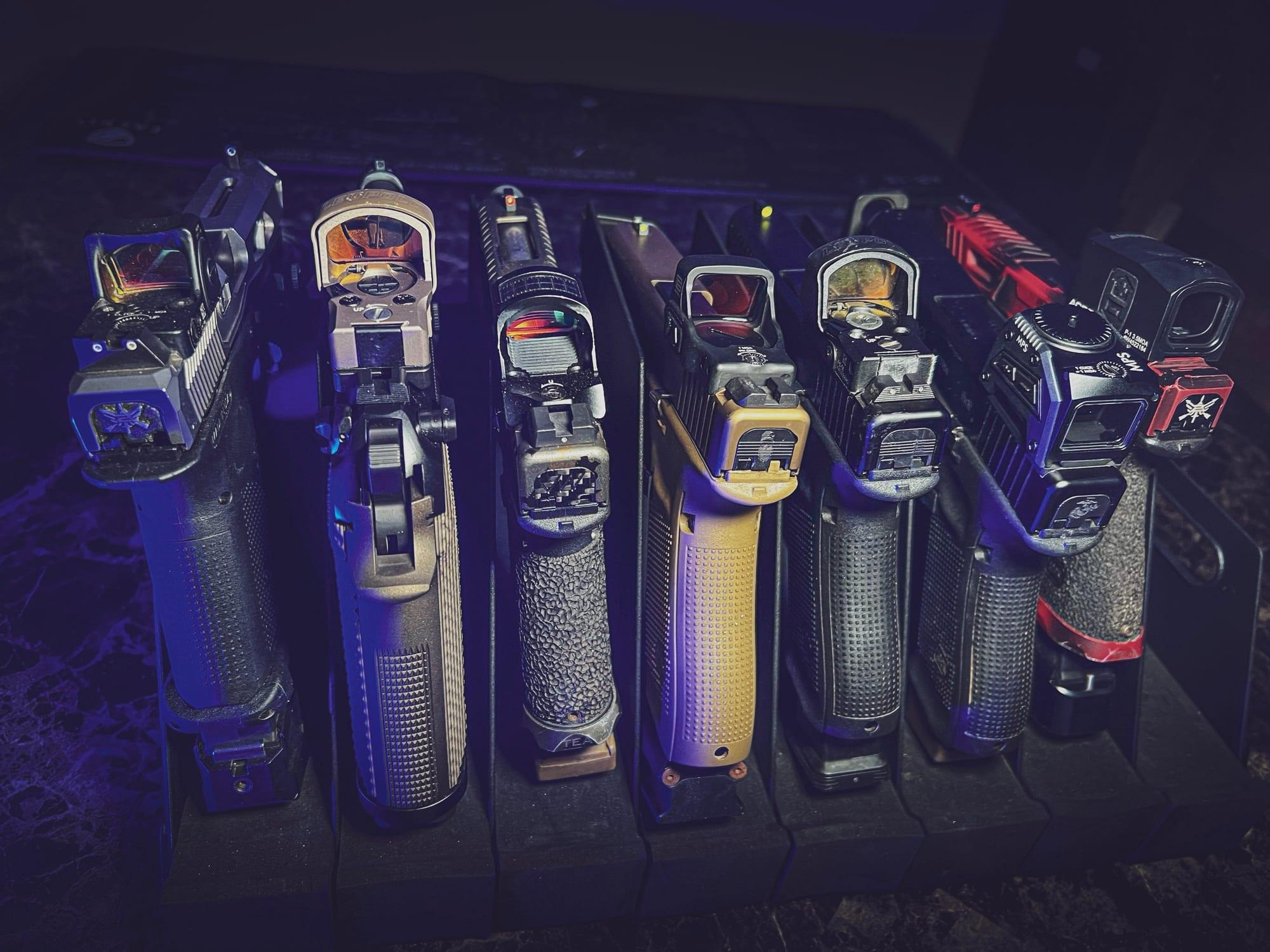 Seven pistols standing muzzle-up in a rack, each wearing a different red dot optic